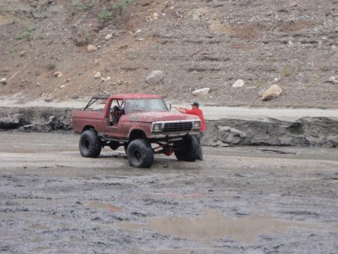 1978 Ford Bronco - A red SUV with large tires navigates a muddy terrain, with a person leaning on the front.