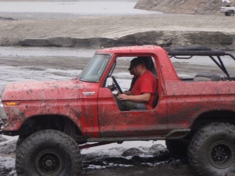 1978 Ford Bronco - A muddy red 1978 Ford Bronco navigates an off-road terrain with a driver inside.