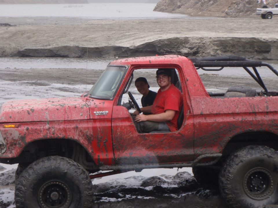Red 1978 Ford Bronco with two occupants, mud-splattered, off-roading