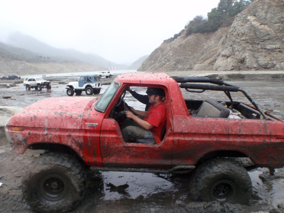 Red 1978 Ford Bronco driving through mud with other off-road vehicles in background