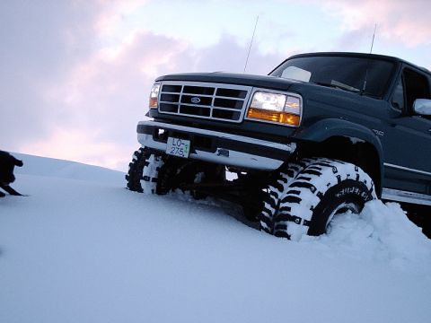 1996 Ford Bronco - A dark green Ford truck navigates deep snow with its large tires partially submerged.