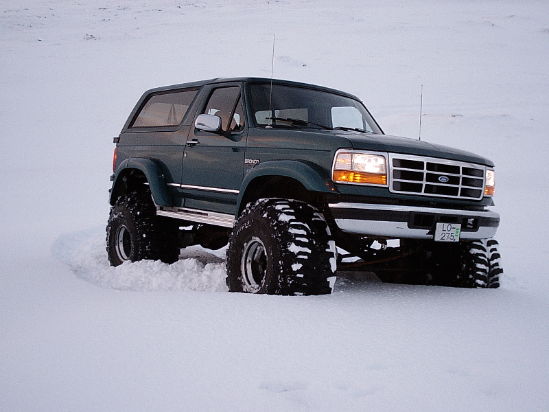 Lifted dark green Ford Bronco on big off-road tires parked in deep snow, front 3/4 view.
