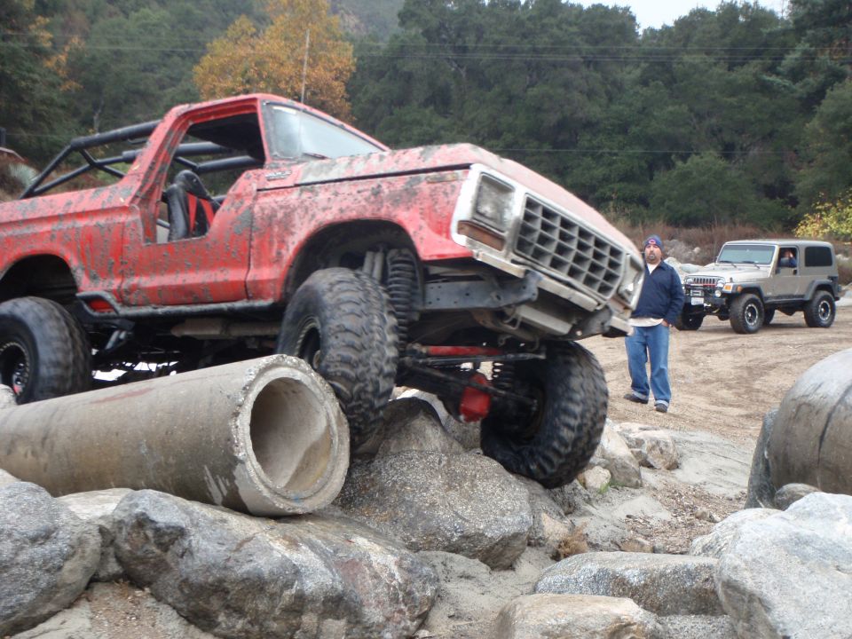Red 1978 Ford Bronco modified for off-roading, climbing over rocks and concrete pipes. Another SUV is parked behind it
