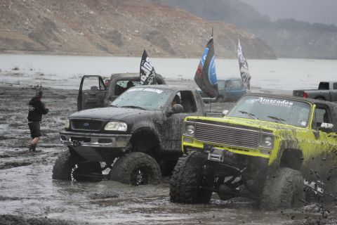 1978 Ford Bronco - A lifted yellow pickup truck navigates through a muddy terrain, with other vehicles and a body of water in the background.