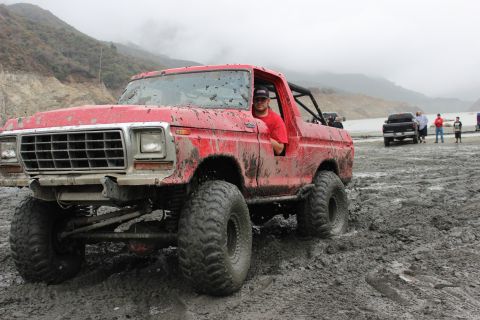 1978 Ford Bronco - A red 1978 Ford Bronco navigates a muddy terrain with other vehicles and people in the background.