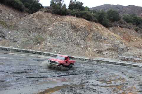 1978 Ford Bronco - A red pickup truck navigates a muddy off-road course with a steep, rocky hillside in the background.