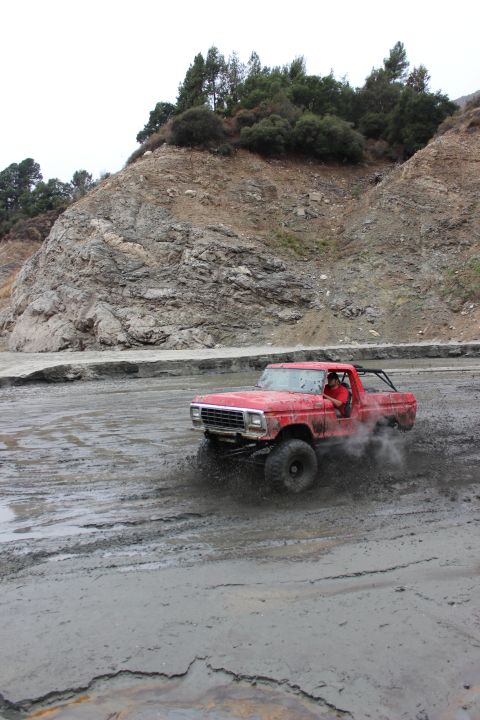 1978 Ford Bronco - A red truck kicks up mud as it drives through a muddy terrain.