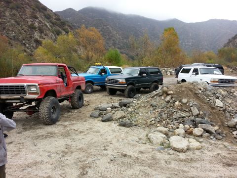 1978 Ford Bronco - A red pickup truck with oversized tires is parked in a rocky, mountainous area alongside other off-road vehicles.