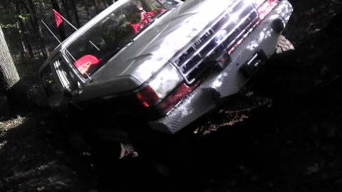 1989 Ford Bronco - A silver 1989 Ford Bronco navigates a wooded trail, its front grille and diamond plate bumper visible.