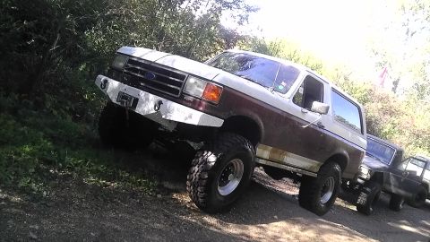 1989 Ford Bronco - Two SUVs, including a 1989 Ford Bronco, are parked on a dirt trail surrounded by trees.