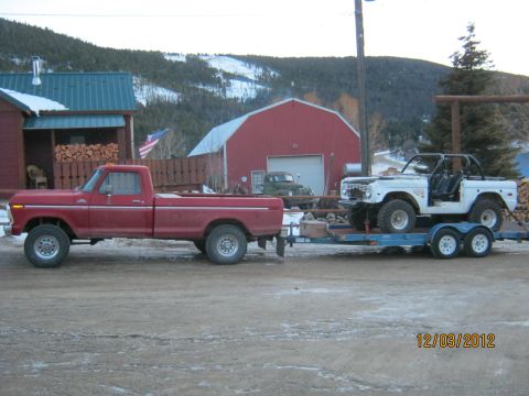1970 Ford Bronco - A red pickup truck tows a white Bronco on a trailer in front of rural buildings and mountains.
