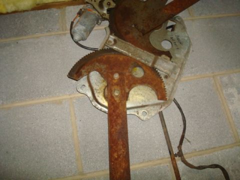 1995 Ford Bronco - A close-up of a heavily rusted window regulator mechanism, likely a car part, is shown against a cinder block background.