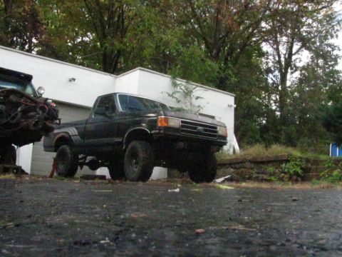 1994 Ford F-150 - A lifted black pickup truck is parked outside a building, with another vehicle's engine in the foreground.