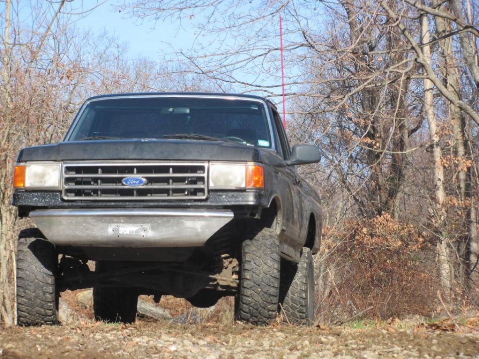 Front view of a lifted black Ford F-150 pickup truck with aftermarket bumper and large tires, parked on a dirt road in a wooded area