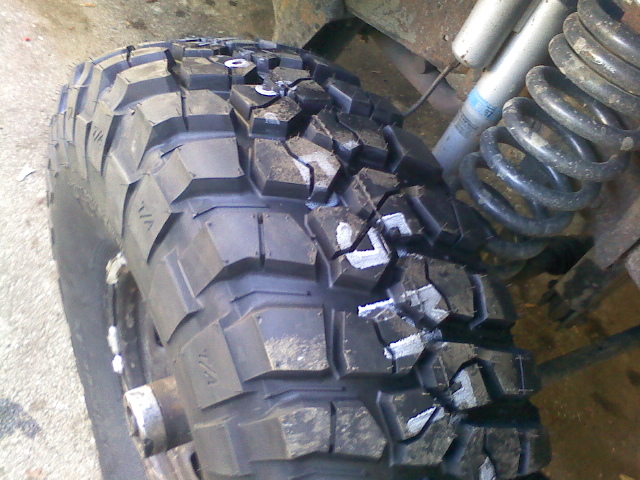 Close-up of a large off-road tire with white markings on the tread. A coil spring and shock absorber are visible in the background