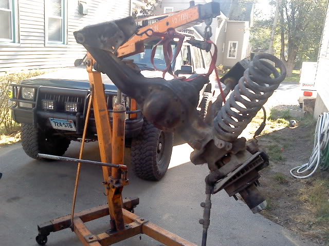 Front axle assembly with coil spring and shock absorber, suspended by an engine hoist over a dark green Jeep Cherokee