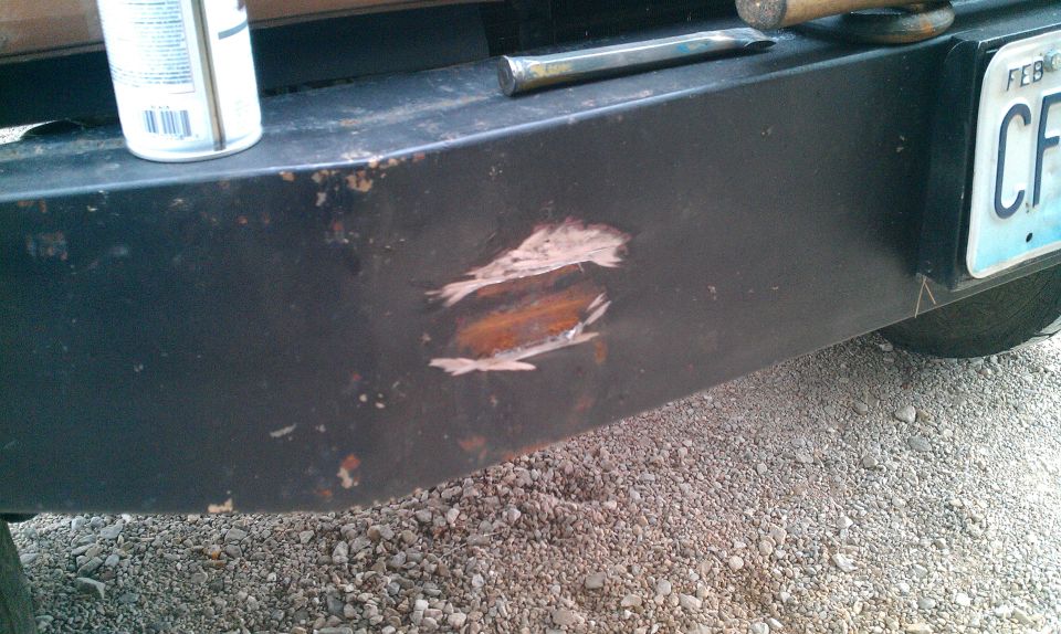 Scratched black bumper with rust showing. License plate visible. Can of spray paint and tool on top
