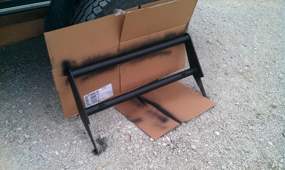 Black push bar being spray painted, resting on cardboard
