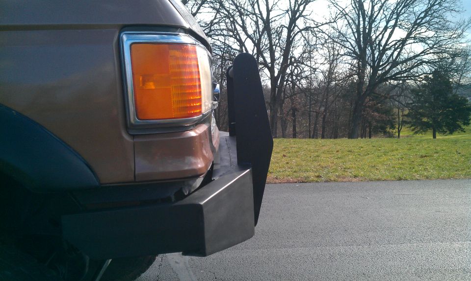 Front corner of a brown SUV with a black push bar and amber turn signal. Trees and a grassy hill in background
