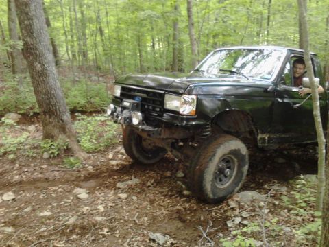 1994 Ford F-150 - A black pickup truck navigates a muddy, wooded path with large off-road tires.