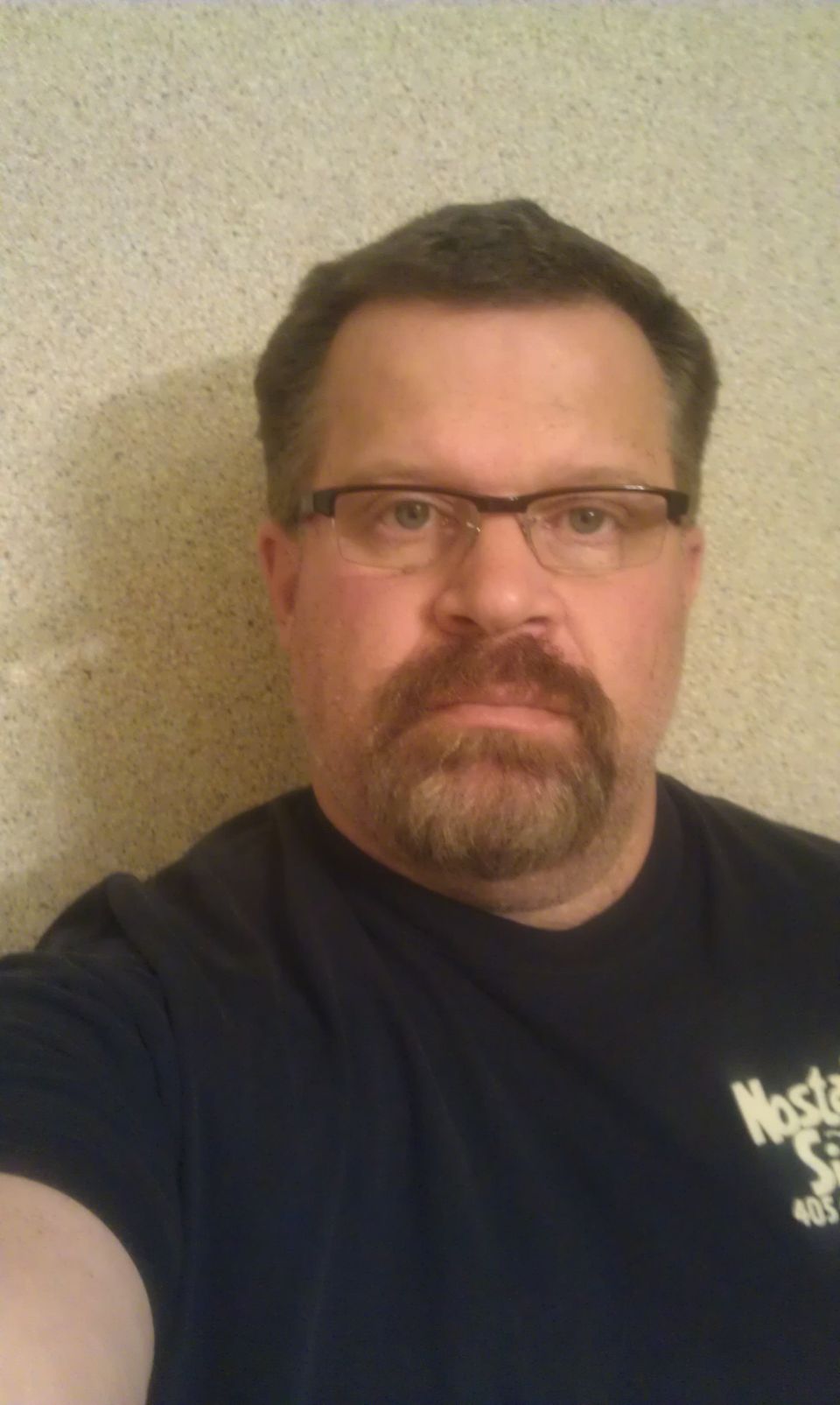 Man with glasses and beard wearing a dark t-shirt with text and a logo