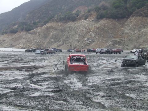 1978 Ford Bronco - Multiple off-road vehicles navigate a muddy terrain near a body of water and rocky hills.