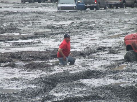 1978 Ford Bronco - A man kneels in thick mud next to a red truck's tire, with other vehicles visible in the background.