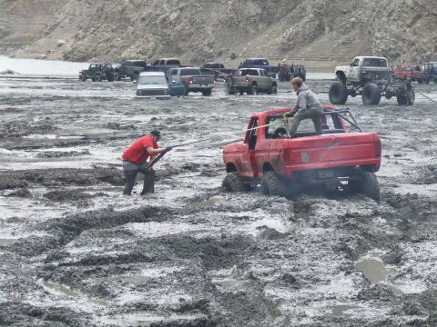 1978 Ford Bronco - A red 1978 Ford Bronco is stuck in thick mud, with two people attempting to free it.