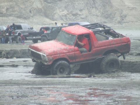 1978 Ford Bronco - A red off-road vehicle is stuck in a muddy area with other vehicles and spectators present.