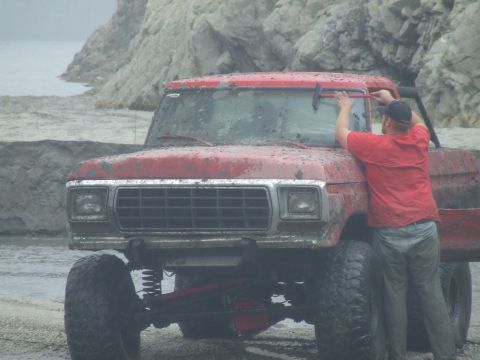 1978 Ford Bronco - A muddy red 1978 Ford Bronco is parked near a rocky shore as a person cleans its windshield.