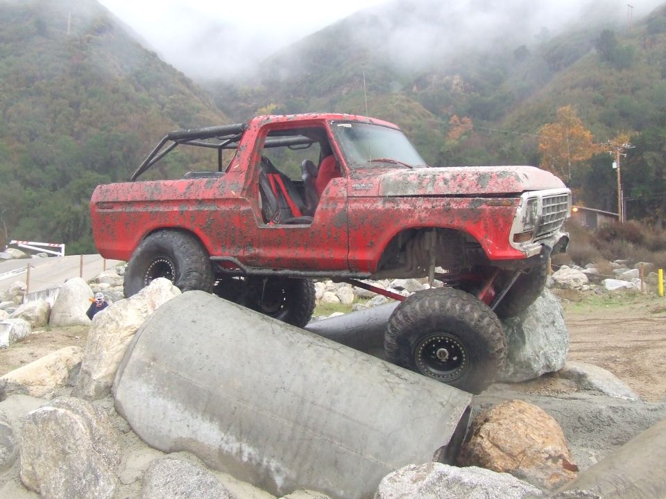 1978 Ford Bronco modified for off-roading, covered in mud, climbing over large rocks and concrete pipes