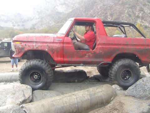 1978 Ford Bronco - A red 1978 Ford Bronco is driven over large rocks and logs off-road.
