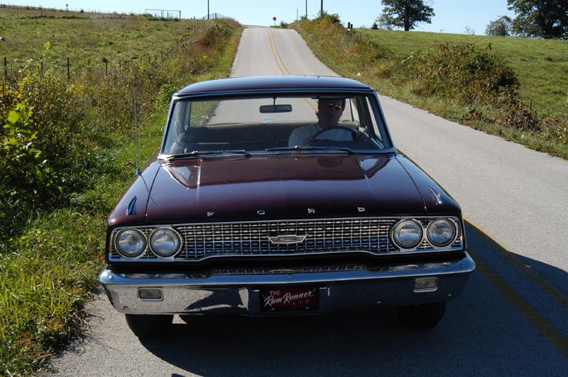 Front view of maroon 1963 Ford Galaxie with 'Rum Runner' plate on a rural country road, driver inside