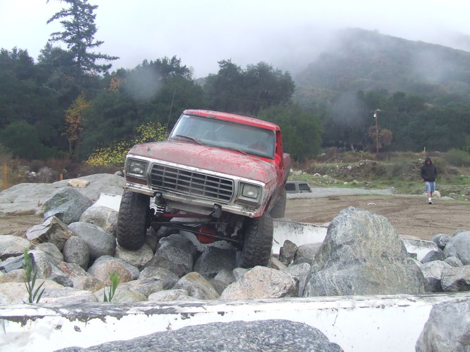 Red pickup truck climbing over rocks on a trail. A person walks in the background