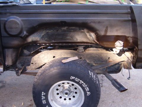 1990 Ford Bronco - The underside of a black truck bed is shown, with the rear wheel and suspension visible. A fuel filler cap is mounted on the side of the be…