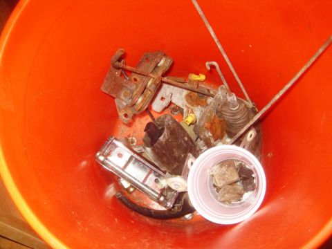 1993 Ford Bronco - Various rusty door latch components are piled inside an orange bucket, with a small plastic cup holding some smaller pieces.