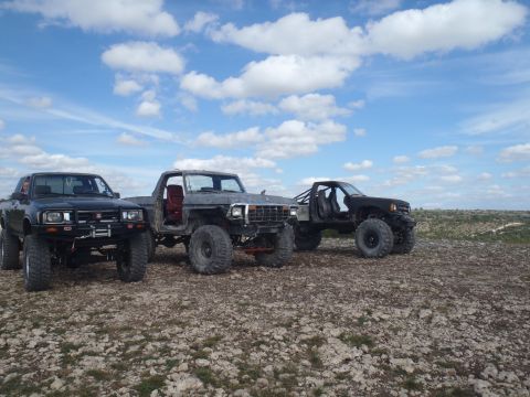 1979 Ford Bronco - Three modified trucks, including a 1979 Ford Bronco, sit parked on a rocky terrain.