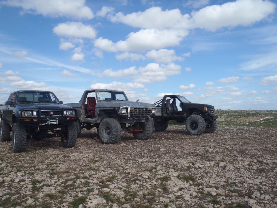 Three modified trucks parked on rocky ground under a cloudy sky. The middle truck appears to be a 1979 Ford Bronco