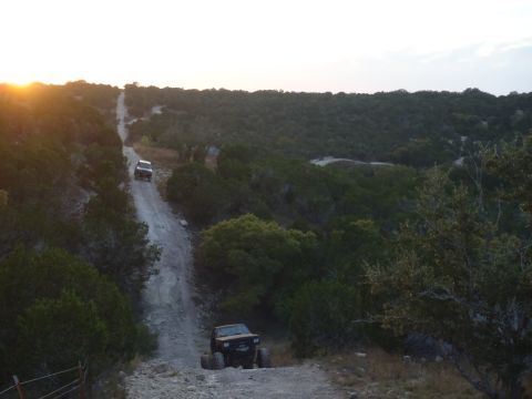 1979 Ford Bronco - Two off-road vehicles navigate a steep dirt road through a wooded landscape as the sun sets.
