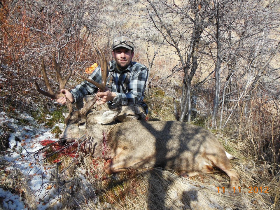 Hunter holding a large buck deer carcass in a wooded area. Watermark reads 'SUPERMOTORS.NET'