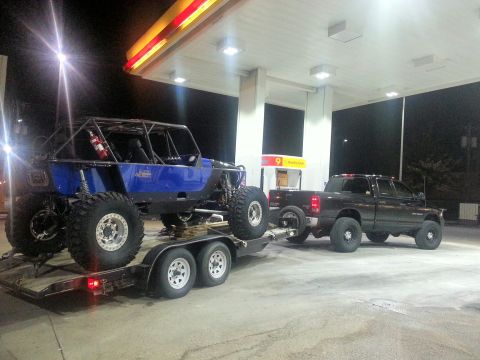 1967 Ford Mustang - A custom blue off-road buggy is transported on a trailer behind a pickup truck at a gas station.