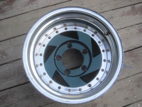 1993 Ford Bronco - A single custom wheel with a chrome lip and teal center is shown against a wooden background. The wheel has multiple bolt holes and a uniqu…
