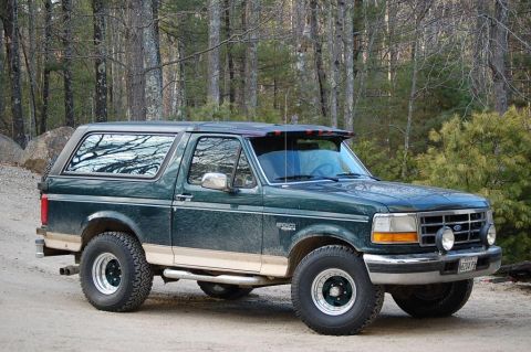 1993 Ford Bronco - A green Ford Bronco is parked on a dirt road with trees in the background.