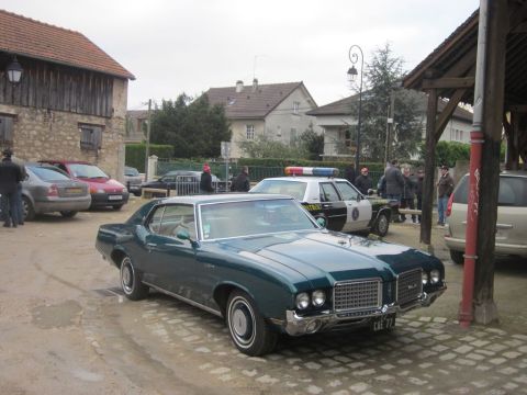 1991 Ford LTD Crown Victoria - A vintage coupe and a police car are parked in a village setting with several people around.