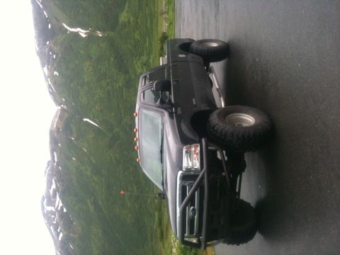 1990 Chevrolet Suburban - A dark-colored pickup truck with large off-road tires and a protective brush guard is parked on a wet road with mountains in the background.
