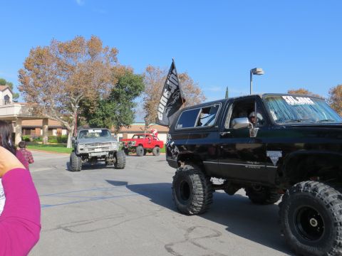 1978 Ford Bronco - Several lifted trucks, including a black SUV with a flag, are seen driving in a parking lot on a sunny day.