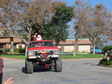 1978 Ford Bronco - Santa drives a lifted 1978 Ford Bronco, waving to onlookers. A blue pickup is parked nearby.