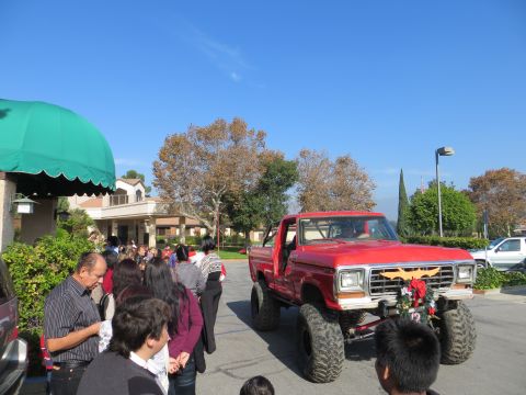 1978 Ford Bronco - A crowd gathers as a lifted red pickup truck, decorated for the holidays, drives by.