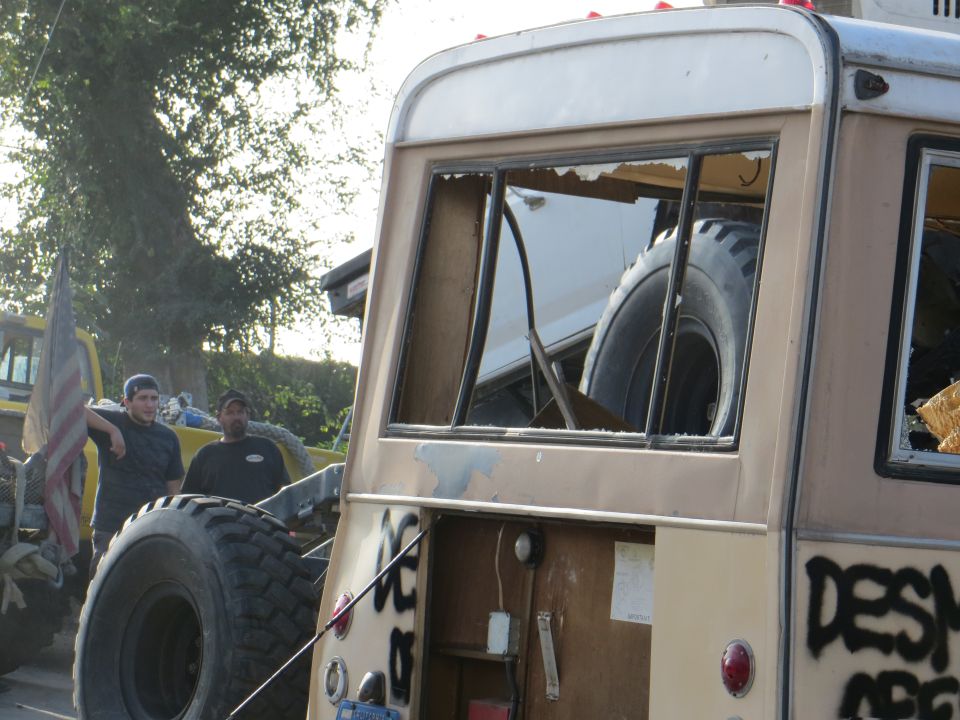 Two men stand near a modified van with large tires and graffiti. A spare tire is visible inside