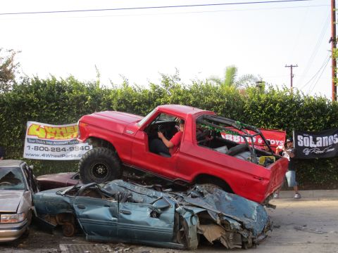 1978 Ford Bronco - A red 1978 Ford Bronco is parked on top of two wrecked cars, with a banner visible.